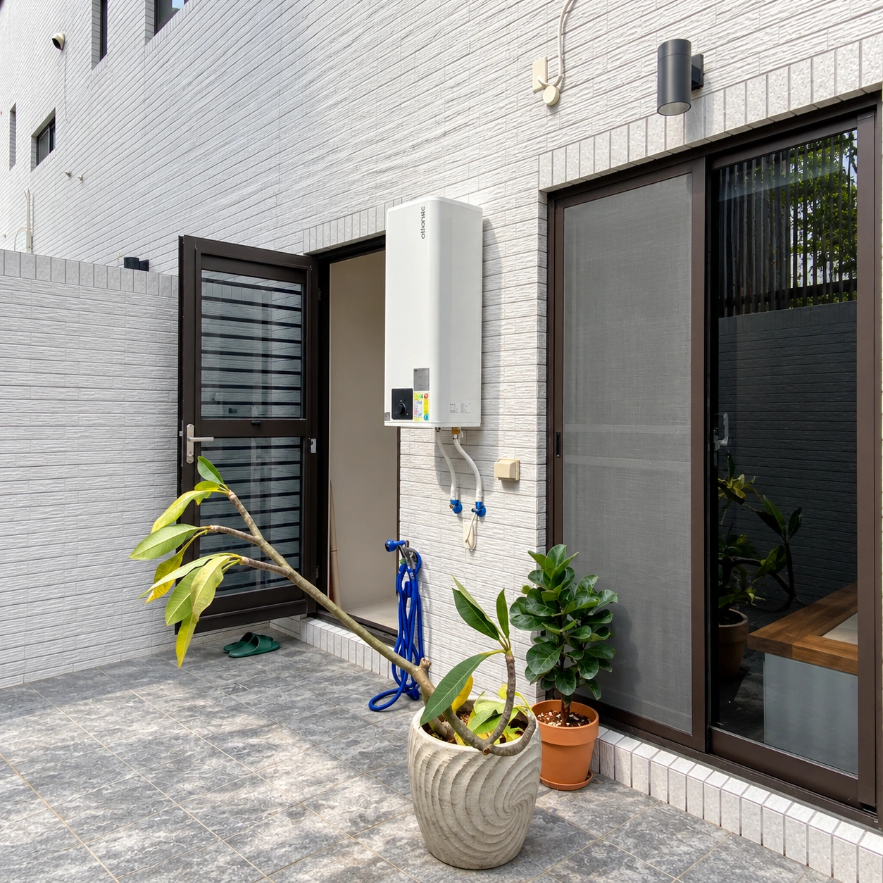 白色直立式熱水器安裝於露臺的白色磚牆，位於黑框門窗旁，下方可見管線、水管與盆栽。