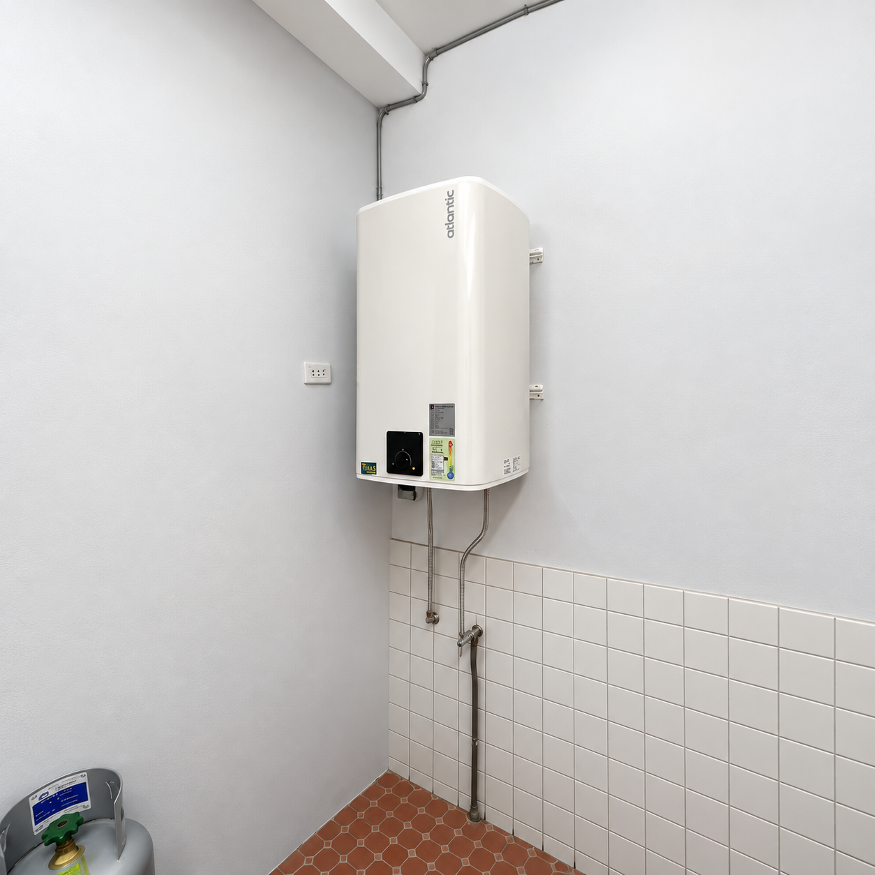 白色方形電熱水器直立壁掛於狹長的磁磚牆邊空間，機身下方可見連接管線與橘色六角地磚。